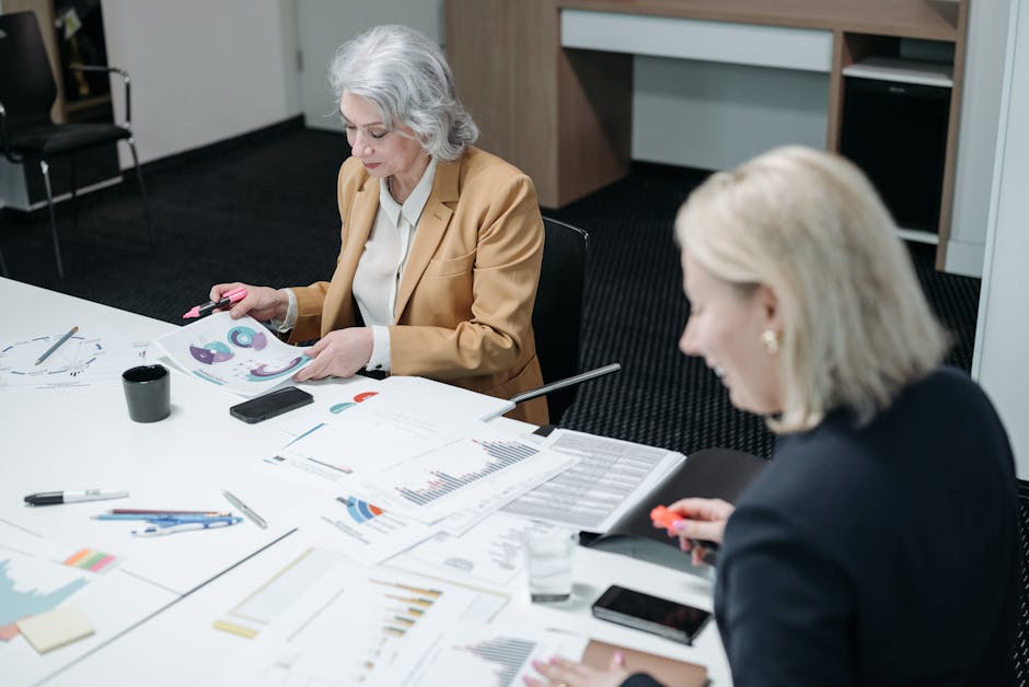 Two senior businesswomen reviewing paperwork and charts in a conference room setting.